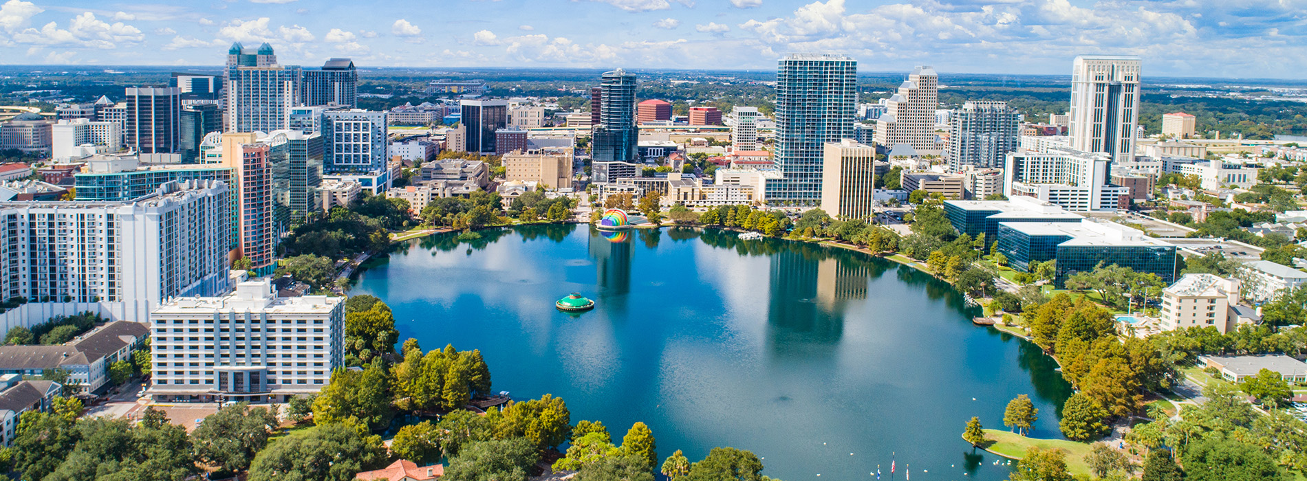 The image shows a city skyline with a large body of water, likely a river or lake, surrounded by buildings and infrastructure, under a clear blue sky with some clouds.