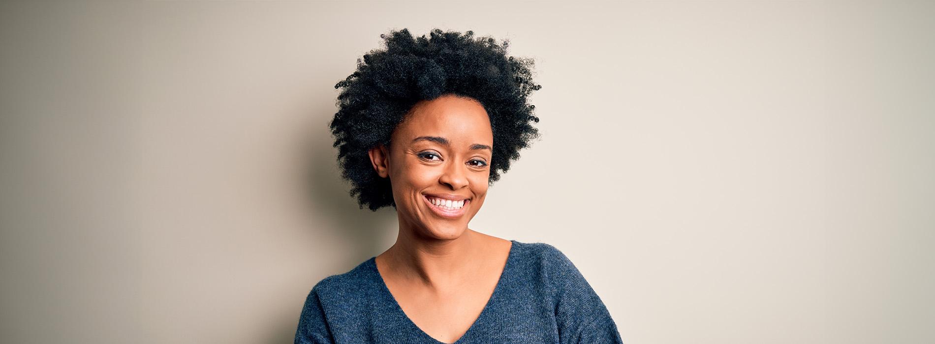 The image is a photograph of a woman with light skin, smiling at the camera. She appears to be in her late twenties or early thirties and has long hair. Her eyes are looking directly at the camera, and she is holding up her index finger near her mouth as if she s making a point or emphasizing something. The background is plain and light-colored, which suggests that this could be a stock photo used for various purposes such as advertising, personal branding, or lifestyle content.