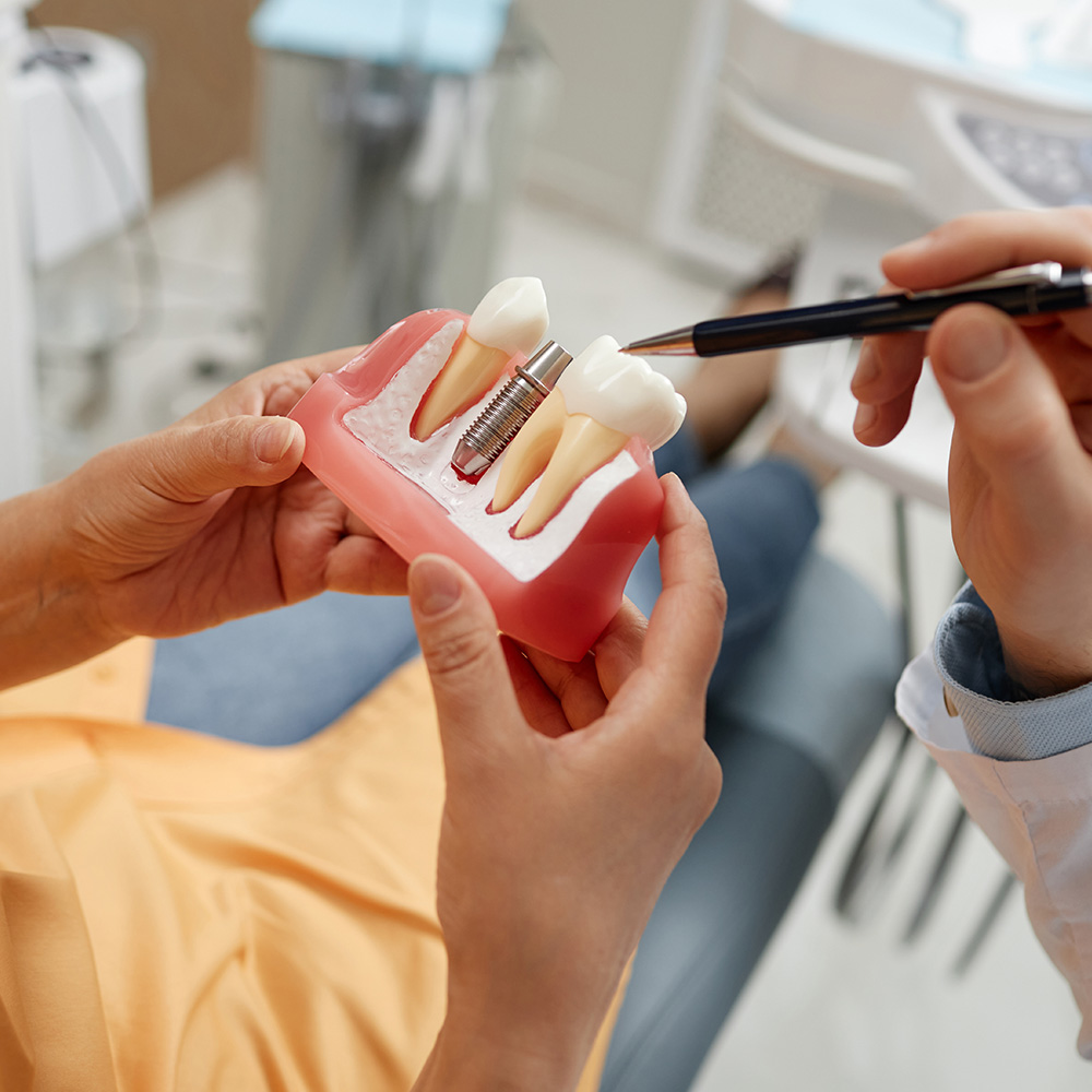A person s hand holding a toothbrush over an open mouth model with fake teeth, set against a dental office background.