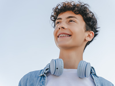 A young person with curly hair wearing headphones, smiling at the camera against a clear sky background.