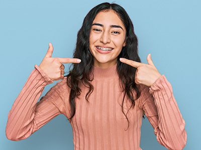 The image shows a woman with long hair pointing at her teeth with both hands, set against a blue background.