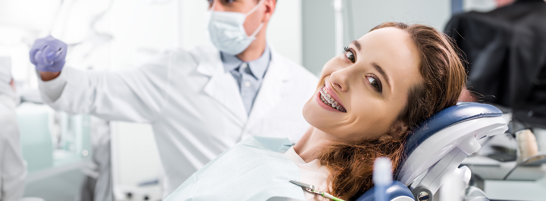 A photograph depicts a woman smiling while seated in a dental chair with a dentist standing beside her, both wearing surgical masks and surrounded by dental equipment.