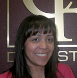 The image shows a woman with short hair smiling at the camera, standing in front of a background with a logo that appears to be related to dental services. She is wearing a pink top and has her hair pulled back.