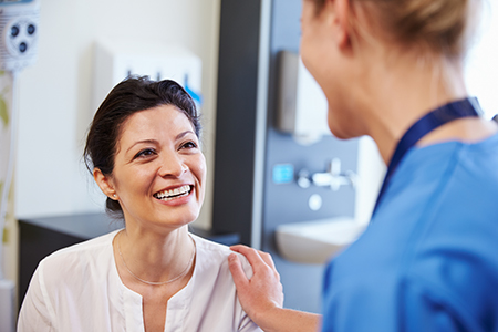 The image shows two people in a medical setting  on the left, there s a woman with a smile, wearing scrubs, standing next to another person who appears to be a healthcare professional, possibly a nurse or doctor, also in scrubs and smiling, with a background that suggests a hospital environment.