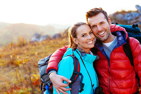 The image shows a man and woman standing outdoors, embracing each other. They are dressed casually with backpacks, suggesting they might be on a hiking trip or outdoor adventure.