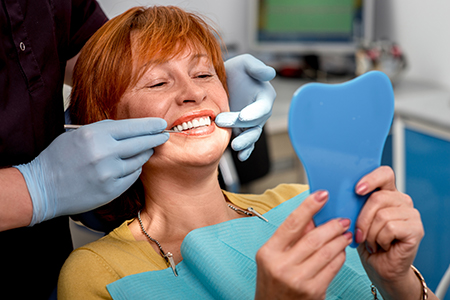 A woman sitting in a dental chair with a blue mouthguard on her teeth, smiling at the camera while looking at a mirror, with a dental professional standing behind her holding the mouthguard up to her mouth.