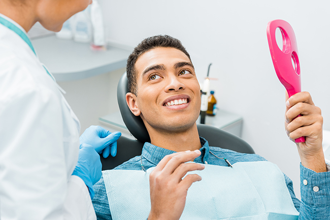 The image shows a person sitting in a dental chair with a smile on their face, holding a pink object, while being attended to by a dental professional who appears to be cleaning teeth.