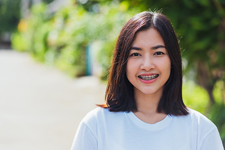 A young woman with short hair smiling at the camera, wearing a white t-shirt and standing outdoors next to a tree.
