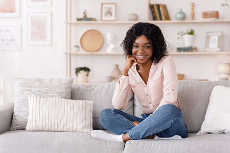 A young woman with curly hair sitting on a couch indoors, smiling at the camera.