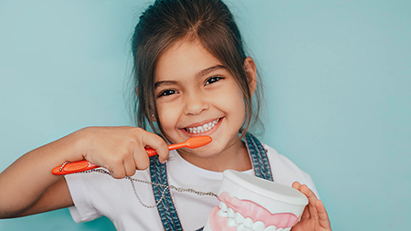 A young girl brushing her teeth with an orange toothbrush while holding a toothpaste tube.