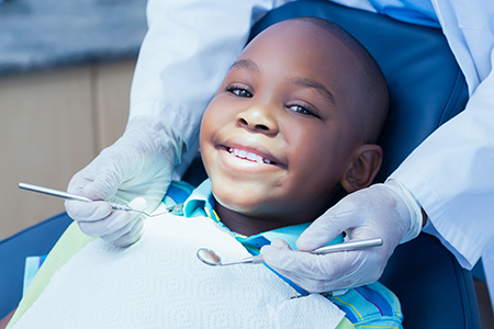 A young boy sitting in a dental chair with a smiling expression, receiving dental treatment from a dentist wearing gloves and a mask.