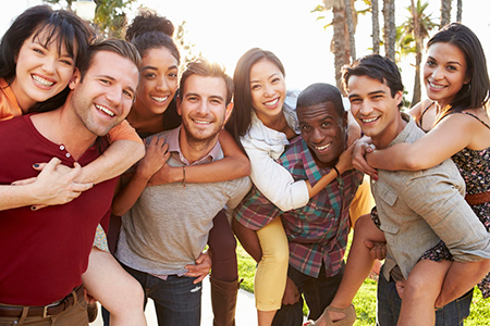 The image shows a group of people posing together outdoors during the daytime, with a warm and sunny atmosphere. They are smiling and appear to be enjoying themselves while hugging each other.