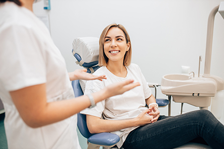 A dental professional is seated at a chair with a patient sitting on it, both wearing face masks, in an office setting.