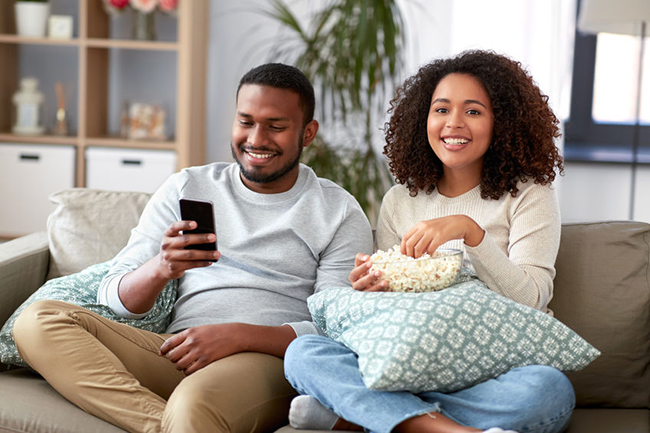 A man and woman sitting on a couch, enjoying popcorn while watching television.