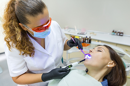 The image shows a dental hygienist performing a teeth cleaning procedure on a patient in a dental office setting.